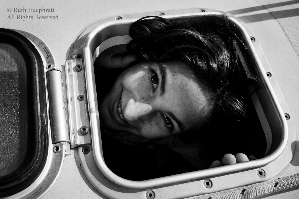 Black-and-white close-up of a smiling woman with freckles looking up through a metal boat hatch