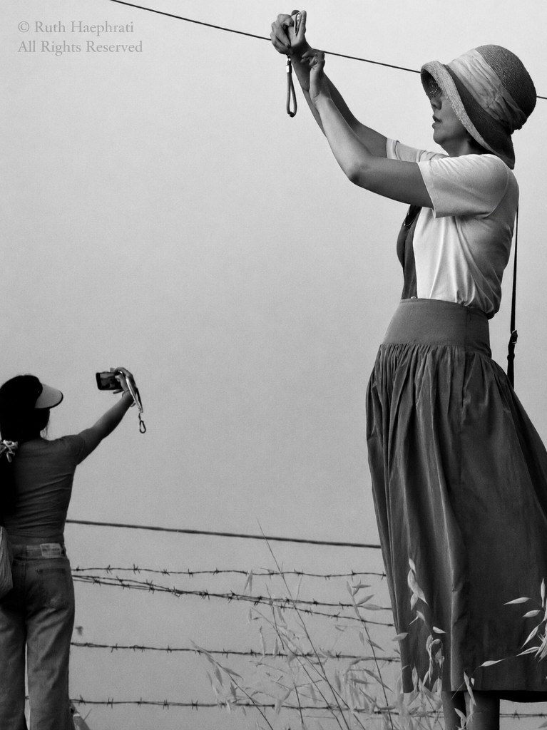 Two people photographing against a bright sky beside a fence, one in a wide-brimmed hat