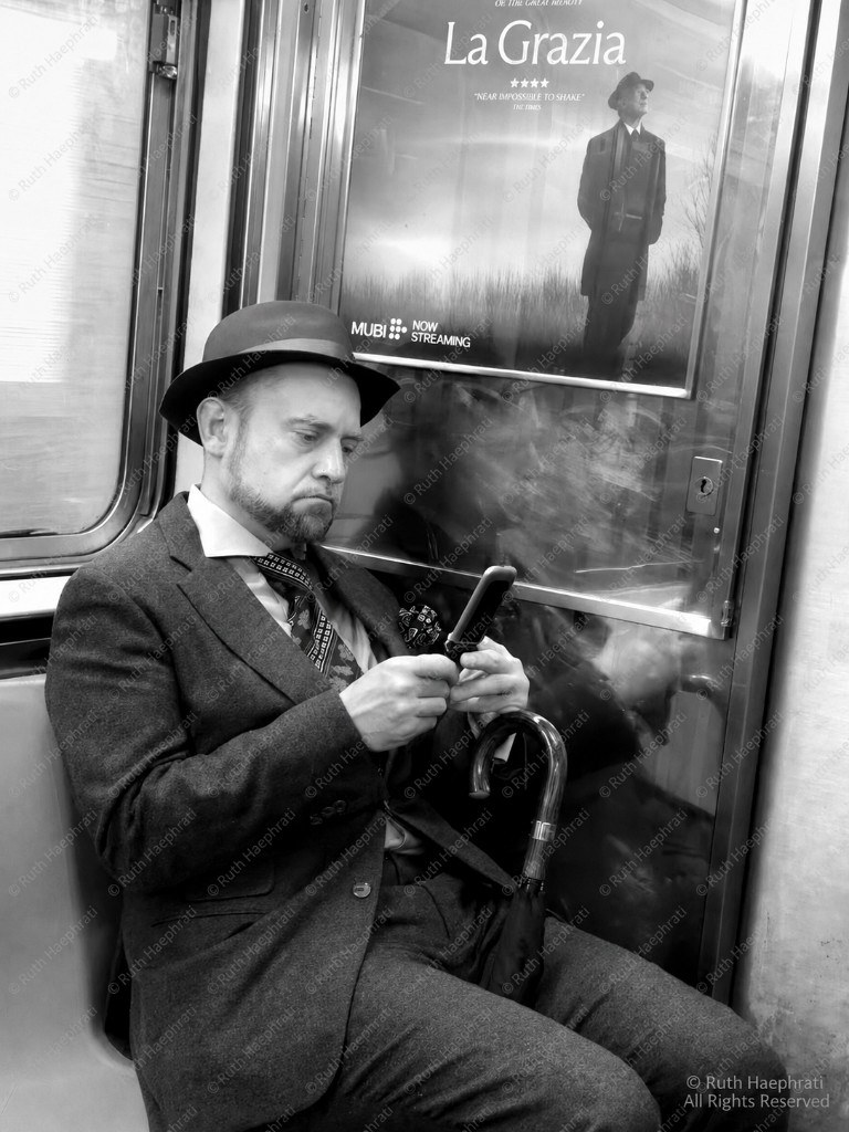Man in a suit and fedora reading a phone on the subway