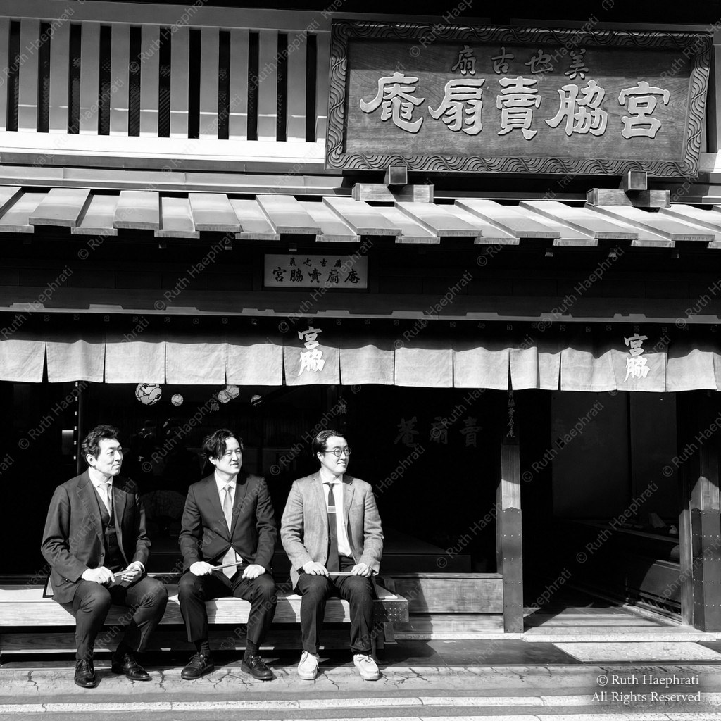 Three men in business suits seated on a bench before a traditional Japanese storefront
